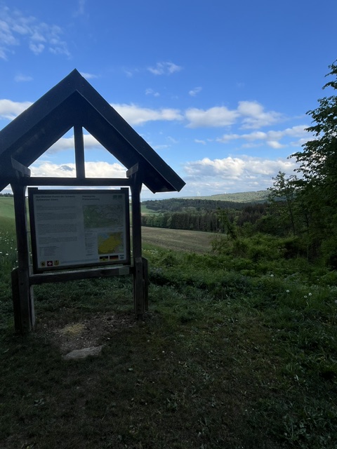 A sign in the shape of a roof with a map on it stands in a field with trees and a blue sky with clouds.